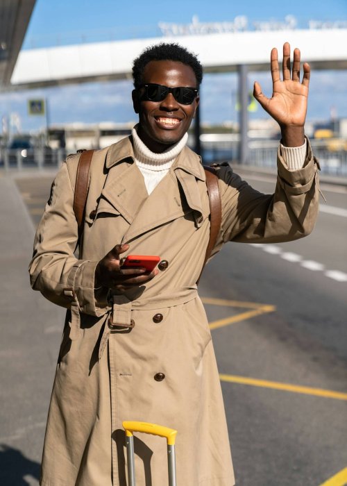 Stylish Black man with suitcase stands in airport, using phone, calling Buffalo Airport taxi, raising hand, greeting